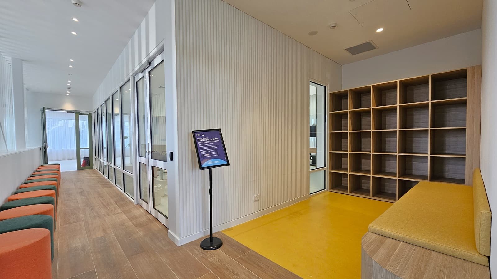 On the left, a row of orange and green cushioned stools lines the wall beneath large windows. In the middle, a corridor with wooden flooring leads to a glass door at the far end, with a sign on a stand in front of a white panelled wall. On the right, a yellow bench is adjacent to a wall of wooden cubby shelves. The floor transitions from wood to yellow vinyl, and the ceiling is white with recessed lighting.