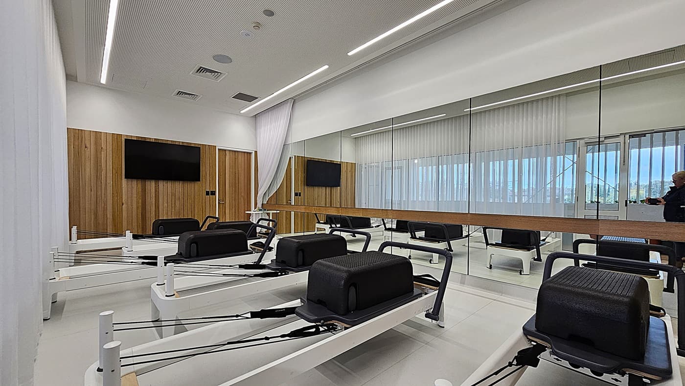 On the left, a wooden panelled wall with a mounted television is next to a closed wooden door. In the middle, several Pilates reformer machines with black cushions and white frames are aligned in rows. On the right, a large wall mirror reflects the room, showing more reformer machines and a person near the entrance. The floor is light-coloured and smooth, and the ceiling is white with recessed lighting and ventilation panels.