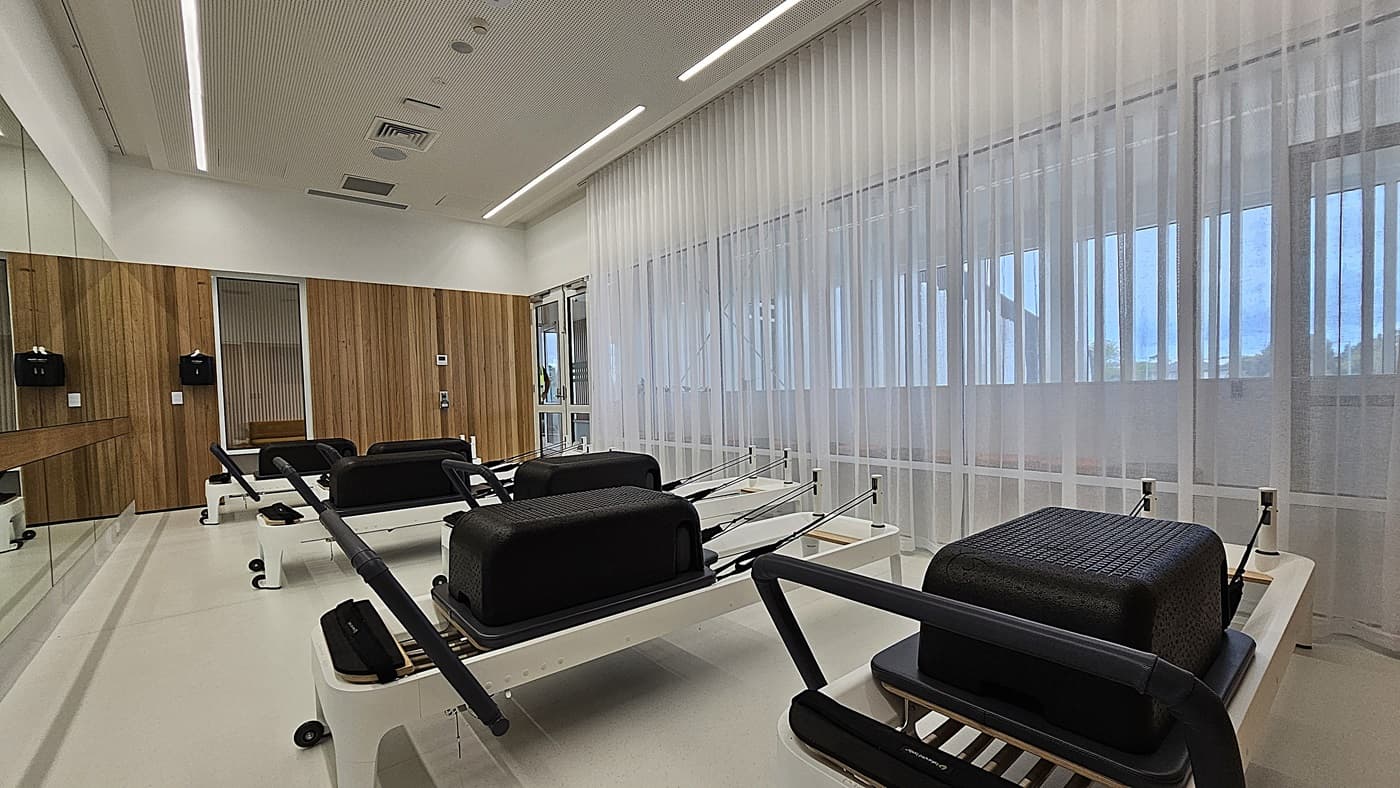 On the left, a wall with vertical wooden panels and a large mirror has two wall-mounted black dispensers. In the middle, several Pilates reformer machines with black cushions and white frames are aligned in rows. On the right, large windows covered with sheer white curtains allow light into the room. The floor is light-coloured and smooth, and the ceiling is white with recessed lighting and ventilation panels.