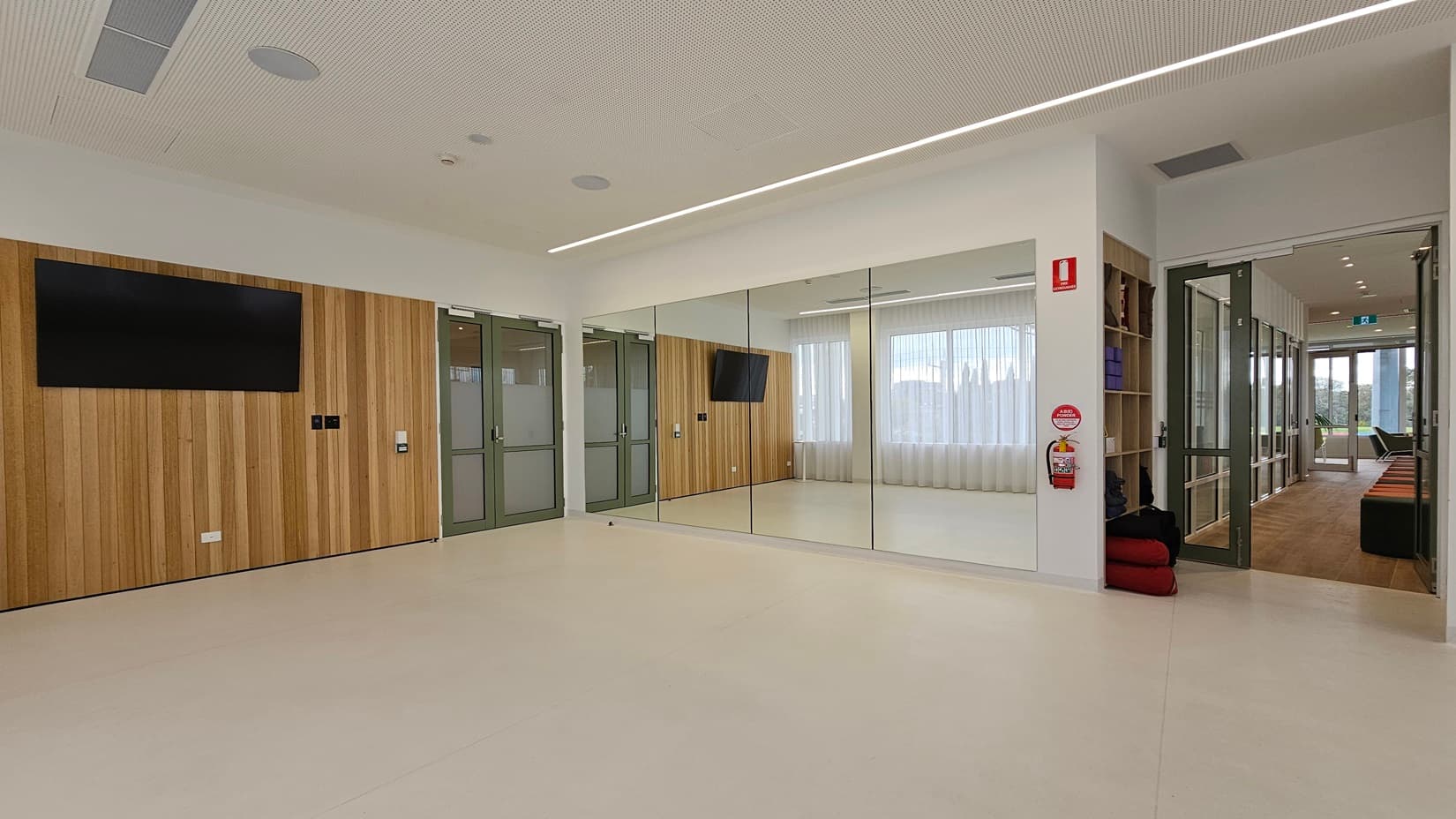 On the left, a wall with vertical wooden panels features a large mounted television and two green double doors with frosted glass panels. In the middle, a large wall mirror reflects the room, showing the wooden panelled wall and windows with sheer white curtains. On the right, an open doorway leads to a corridor with wooden flooring and seating. The floor is light-coloured and smooth, and the ceiling is white with recessed lighting and ventilation panels.