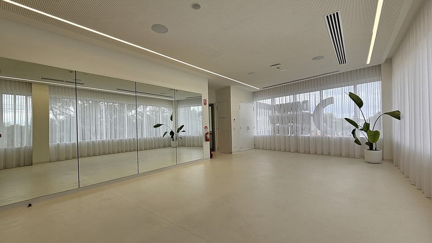 On the left, a wall with large floor-to-ceiling mirrors reflects the room. In the middle, a white column with a fire extinguisher mounted on it stands near a closed door. On the right, large windows with sheer white curtains allow natural light to fill the space, and a potted plant with broad green leaves is placed near the corner. The floor is light-coloured and smooth, and the ceiling is white with recessed lighting and ventilation panels.