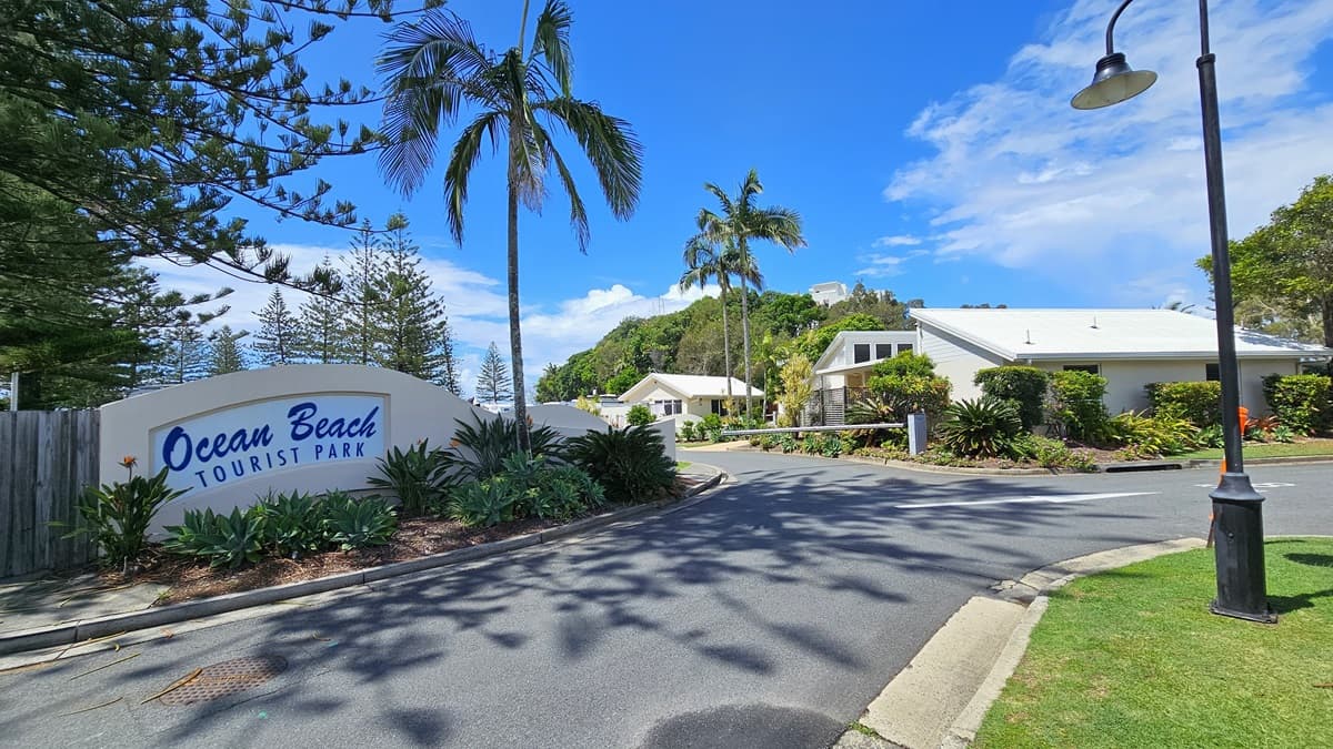 To the left, a concrete landmark displays the name "Ocean Beach Tourist Park" amid landscaped plants. The bitumen road splits, with a traffic barrier indicating no entry on the left. White markings guide travellers on the right path. Landscaped green grass fills the right side. Lamp posts and palm trees line the road. Low-set buildings with pitched roofs are positioned on the left side of the road.