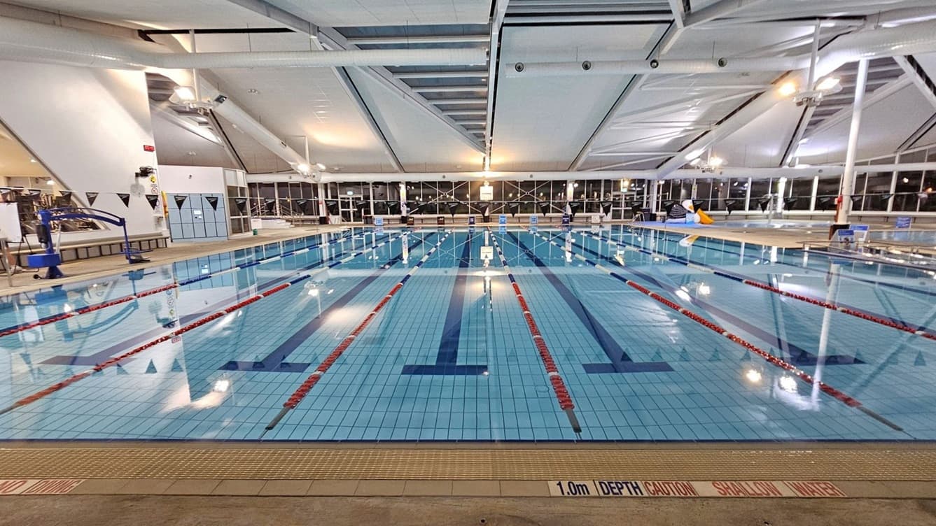 An indoor swimming pool with multiple lanes marked by red and white lane dividers. The pool's floor is tiled in light blue with dark blue lane markings. On the left, there is a pool lift and a person standing near the edge. The right side features a large inflatable penguin. The ceiling is high with exposed beams and lighting fixtures. Large windows line the back wall, allowing natural light to enter. The pool deck is tiled, and a sign indicates a depth of 1.0m.