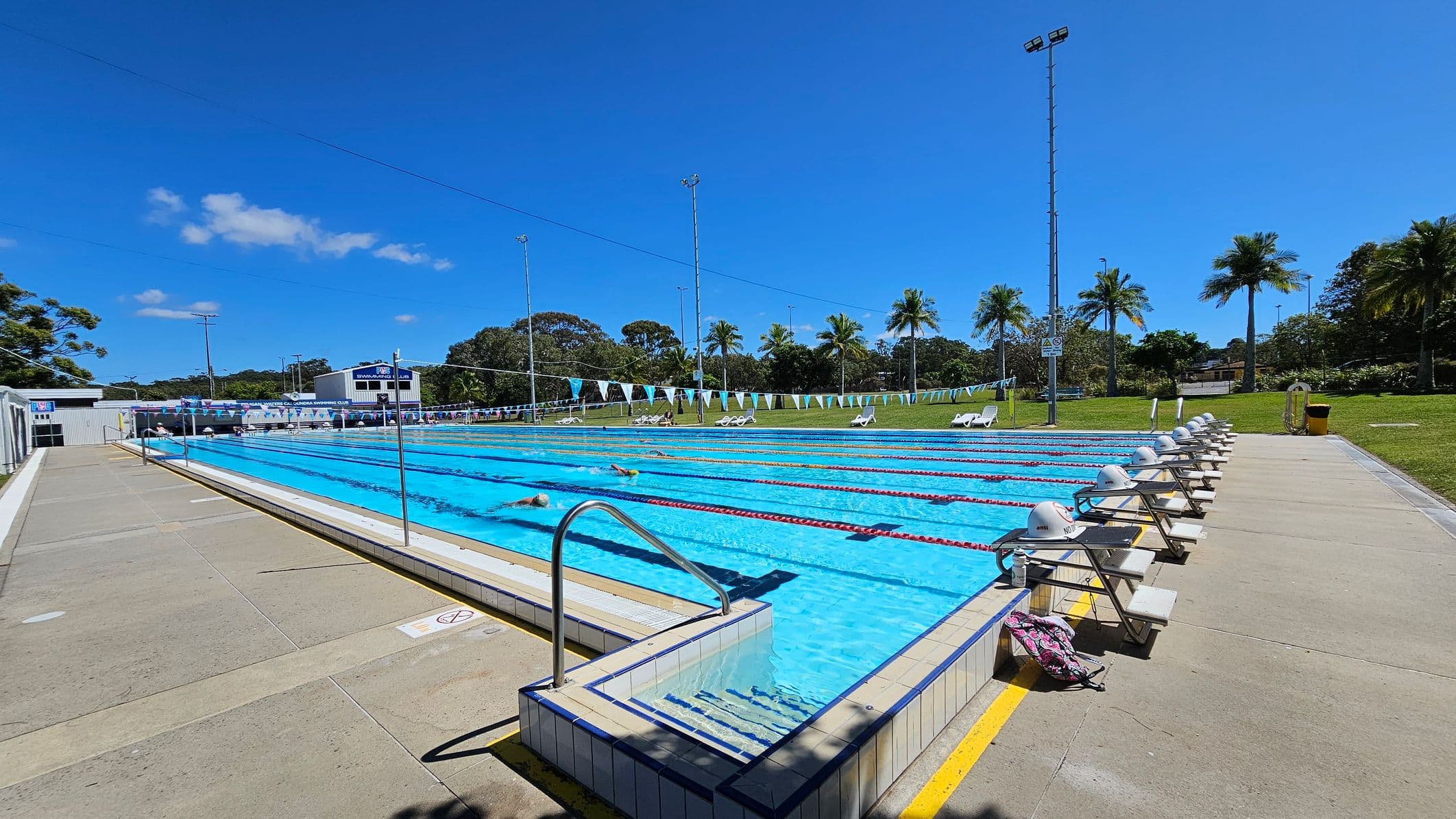 An outdoor swimming pool with multiple lanes, each marked by lane ropes. Starting blocks are positioned at the near end of the pool. On the left, there is a grassy area with trees and a shaded seating area. On the right, there are white lounge chairs on a grassy strip. In the background, there are buildings, including a structure with a sign that reads "Swim Club," and tall light poles surrounding the pool area. The pool deck is concrete.