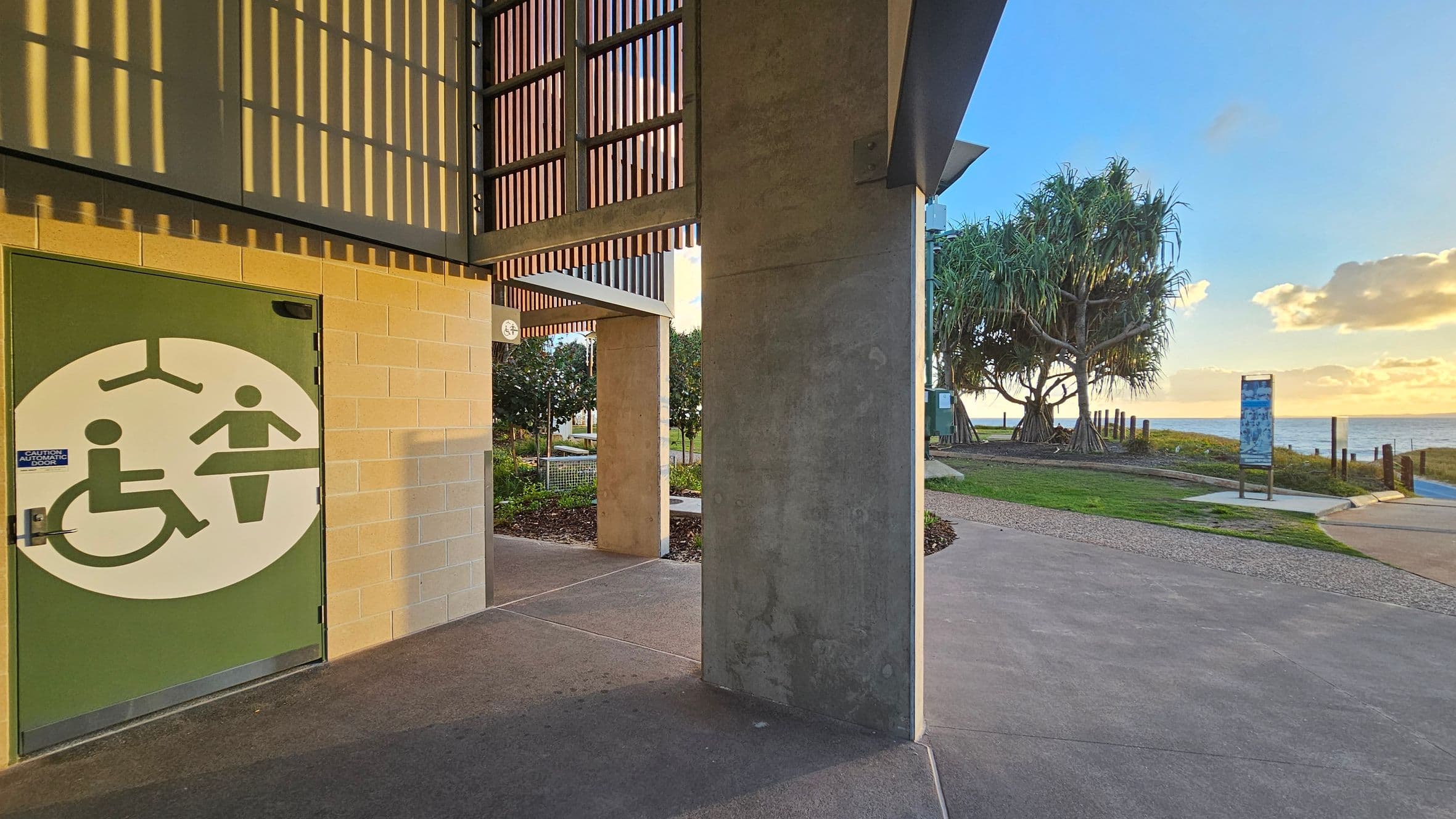 A modern building with vertical slats on the left, featuring a covered walkway. In front, a sign reads "Bluey and Jean Piva Park." The path leads to the right, bordered by well-maintained greenery and palm trees. A solar-powered light pole is visible along the path, which extends towards the horizon. The ground is paved with a mix of concrete and decorative patterns.