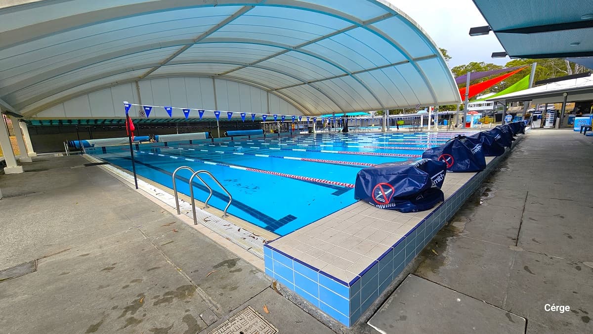 The image shows the Nerang Aquatic Centre's outdoor pool area, featuring a 25m, 10-lane pool covered by a white dome on the left side. There are red and light green shade sails protecting the splash park and toddler wading pool. Water features include a red mushroom fountain, a large blue frog water slide, a light blue mushroom fountain, and a cone bucket area with red, blue, and yellow colors. At the back, there are two large blue tents and a 25m, 4-lane pool separated by a black fence.
