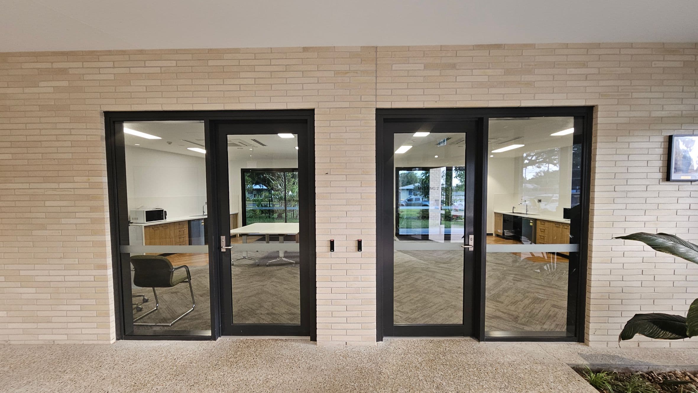 Two glass doors with black frames are set in a light brick wall. The left door reveals a room with a chair and a table, while the right door shows a kitchenette with wooden cabinets and a sink. The floor is speckled concrete, and a large plant is partially visible on the right. Reflections in the glass show outdoor greenery and a parked car.