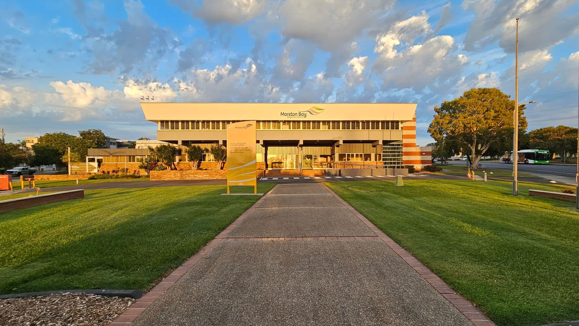 A modern building with a flat roof and large windows is in the centre, with "Moreton Bay" signage on the facade. A wide pathway leads up to the entrance, flanked by well-maintained green lawns. On the right, a large tree and a flagpole are visible, while a bus is seen on the road. On the left, there are smaller trees and a parked vehicle. The sky is partly cloudy, casting a warm light on the scene.
