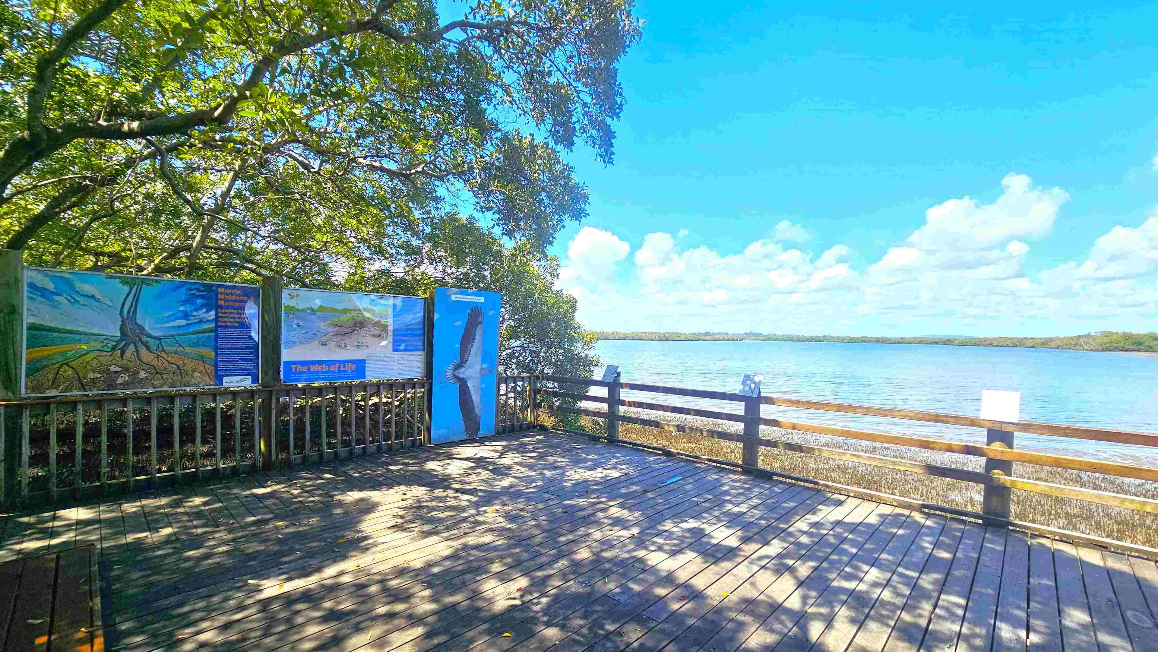 A wooden viewing platform with railings overlooks a body of water surrounded by trees. On the right, there are informational signs featuring images of birds. The platform is partially shaded by overhanging branches. The floor is made of wooden planks, and the background shows a clear view of the water and distant tree line.