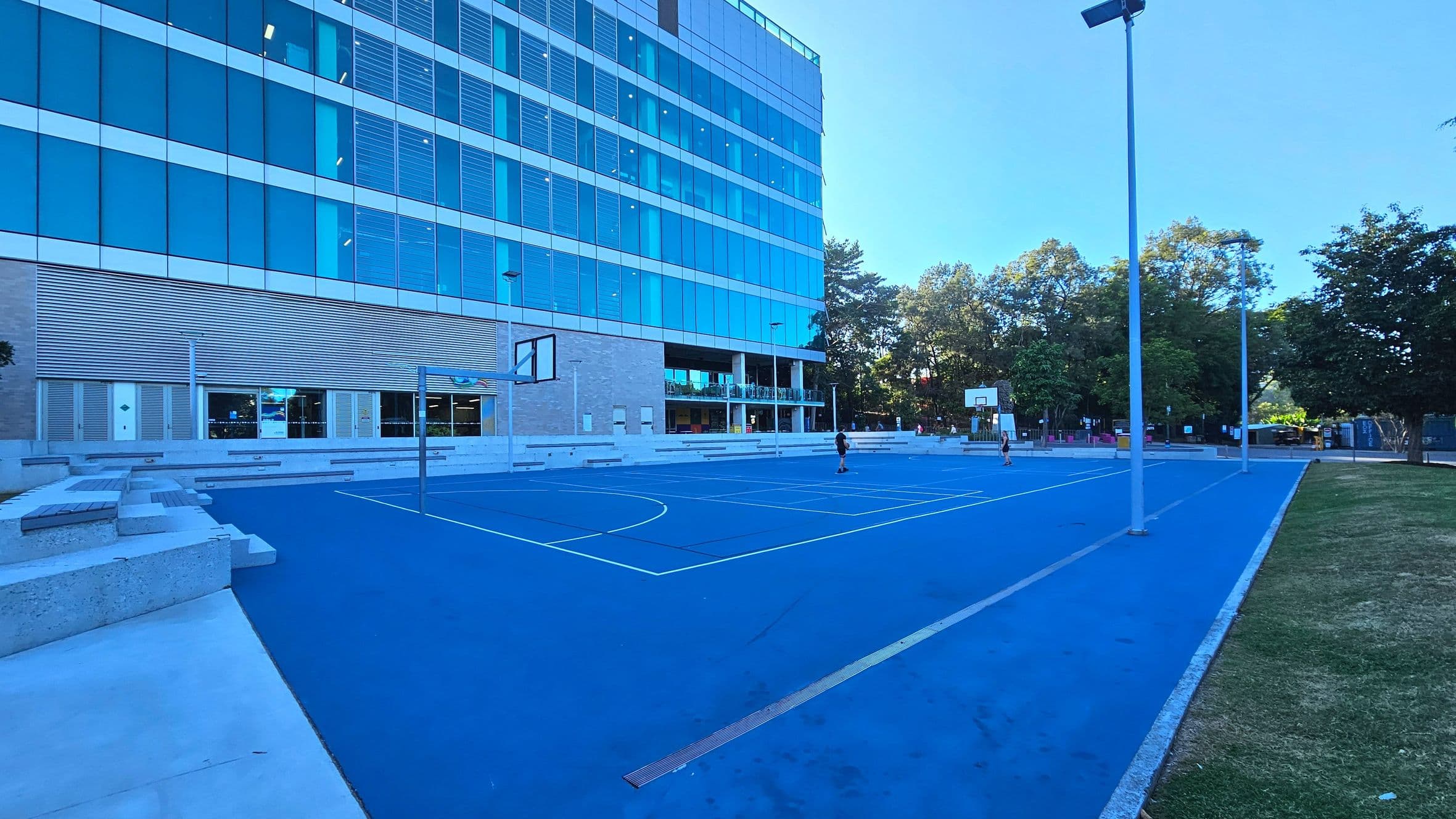 A modern outdoor basketball court with a bright blue surface is adjacent to a multi-storey building with large glass windows and horizontal louvres. The court features two basketball hoops and is surrounded by tall street lamps. Concrete seating lines the edge of the court, and a grassy area borders one side. Trees are visible in the background, providing a natural boundary.