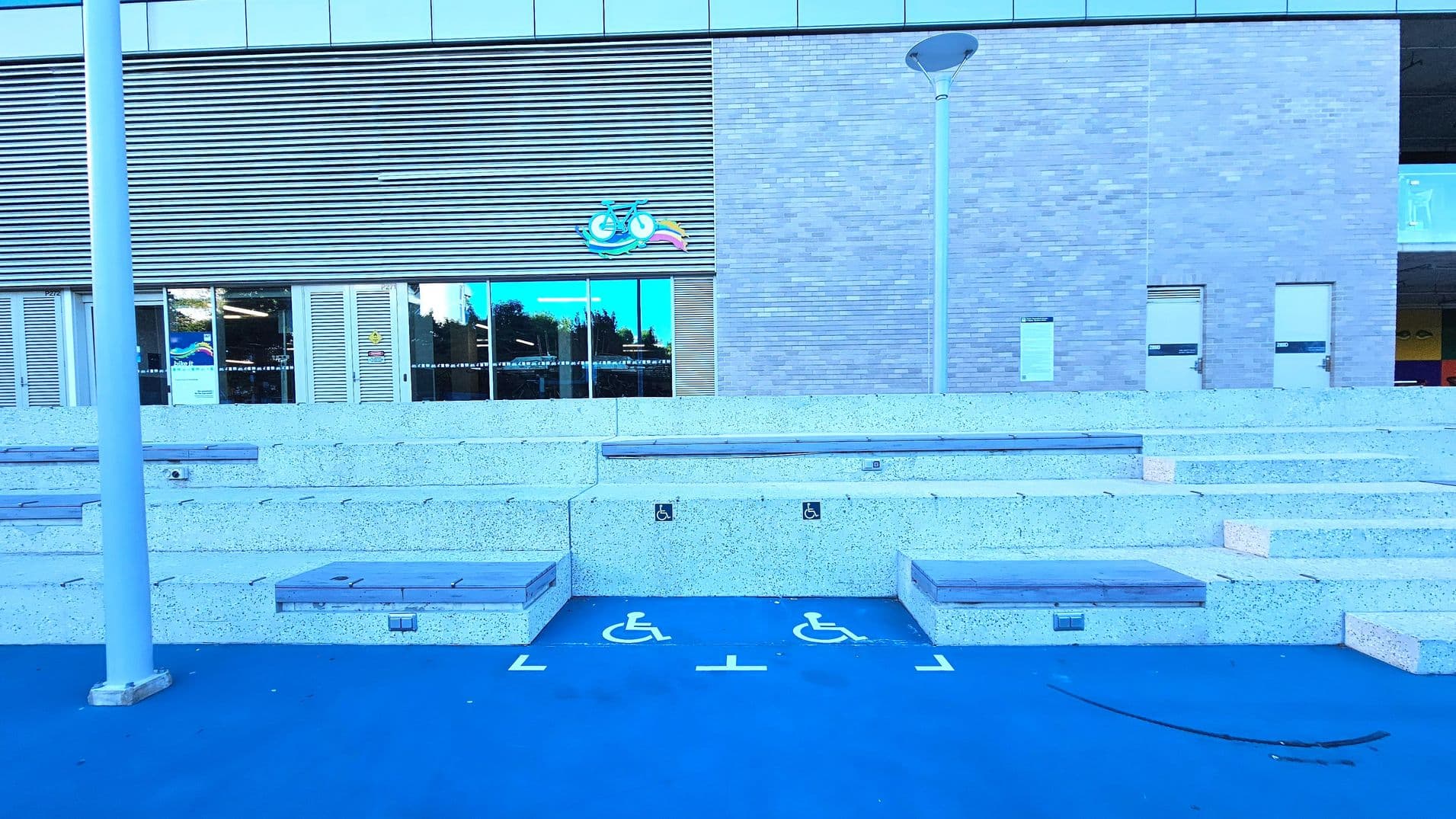 The image shows a modern building facade with a combination of brick and metal louvre panels. In front, there are tiered concrete seating steps with wooden tops, flanked by two designated wheelchair spaces marked on a blue surface. A tall lamp post stands to the left. The building features large glass doors and windows, with a colourful logo above. The area feels open with no visible barriers.