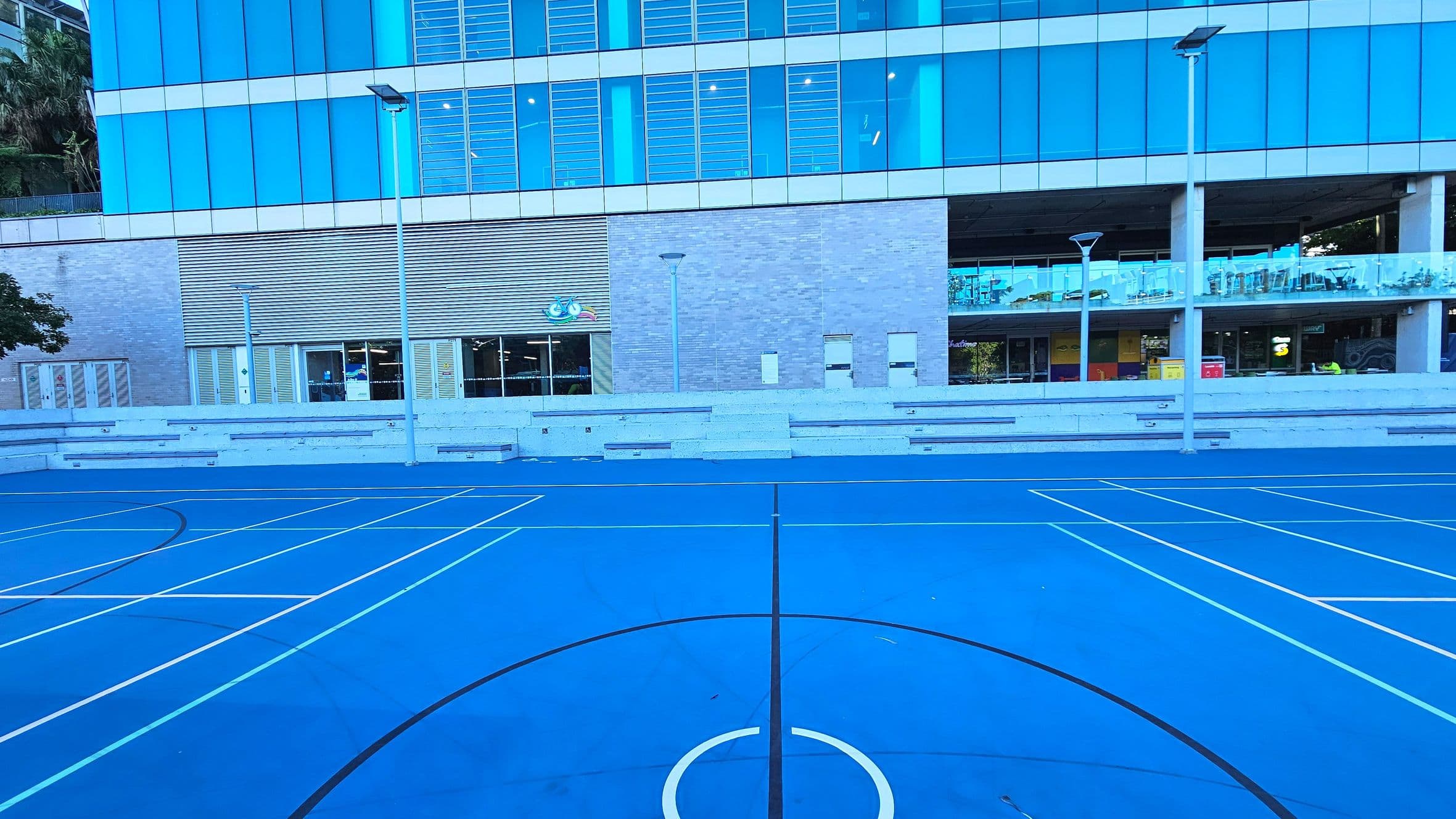 A vibrant blue sports court with white lines for various games is in the foreground. Behind it, a modern building with large blue-tinted glass windows and a grey brick facade stands. The building features a covered walkway with glass railings and several entrances. Tall street lamps line the court, and tiered concrete seating is visible along the building's base.