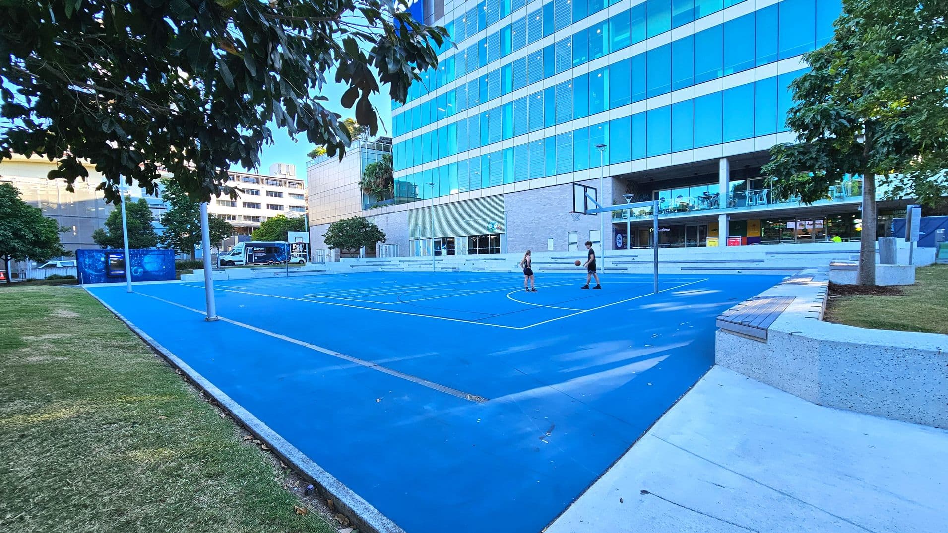 A vibrant blue basketball court is situated next to a modern glass building with reflective windows. Two people are playing basketball on the court, which is surrounded by a low concrete wall with wooden seating. Trees with lush green foliage border the court, and a grassy area is visible to the left. The building features a ground-level entrance and a balcony with seating, suggesting a recreational or commercial space.