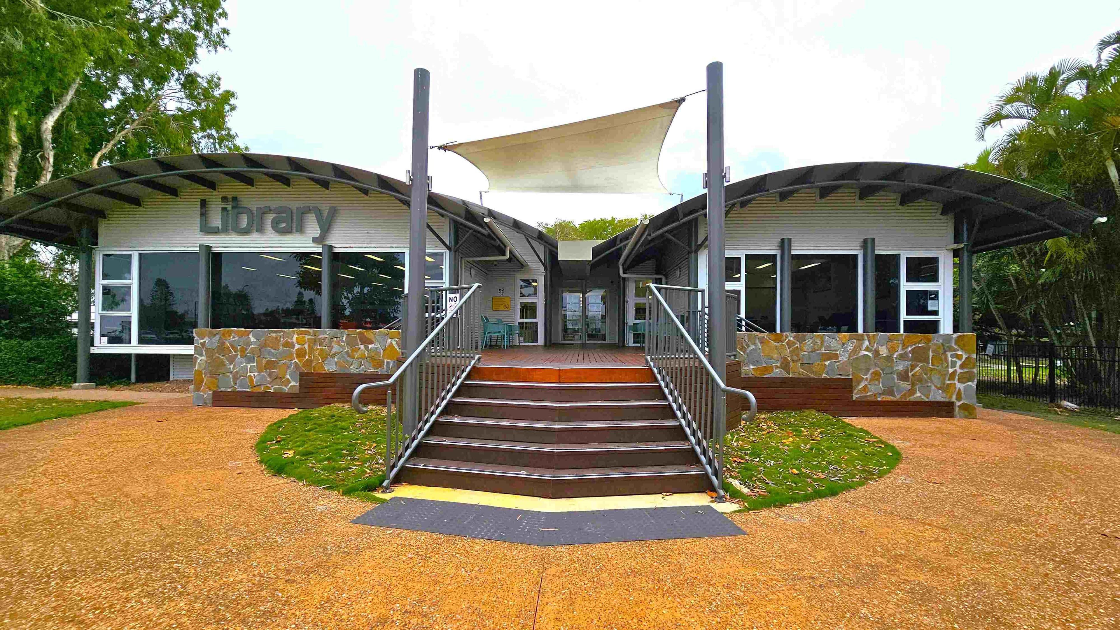 A modern library building with a curved roof and large windows. The entrance is in the centre, accessible by a set of stairs with metal railings. The facade features stonework and a sign reading "Library" on the left. A white shade sail is suspended above the entrance. The ground is a mix of gravel and grass, with trees visible on both sides.