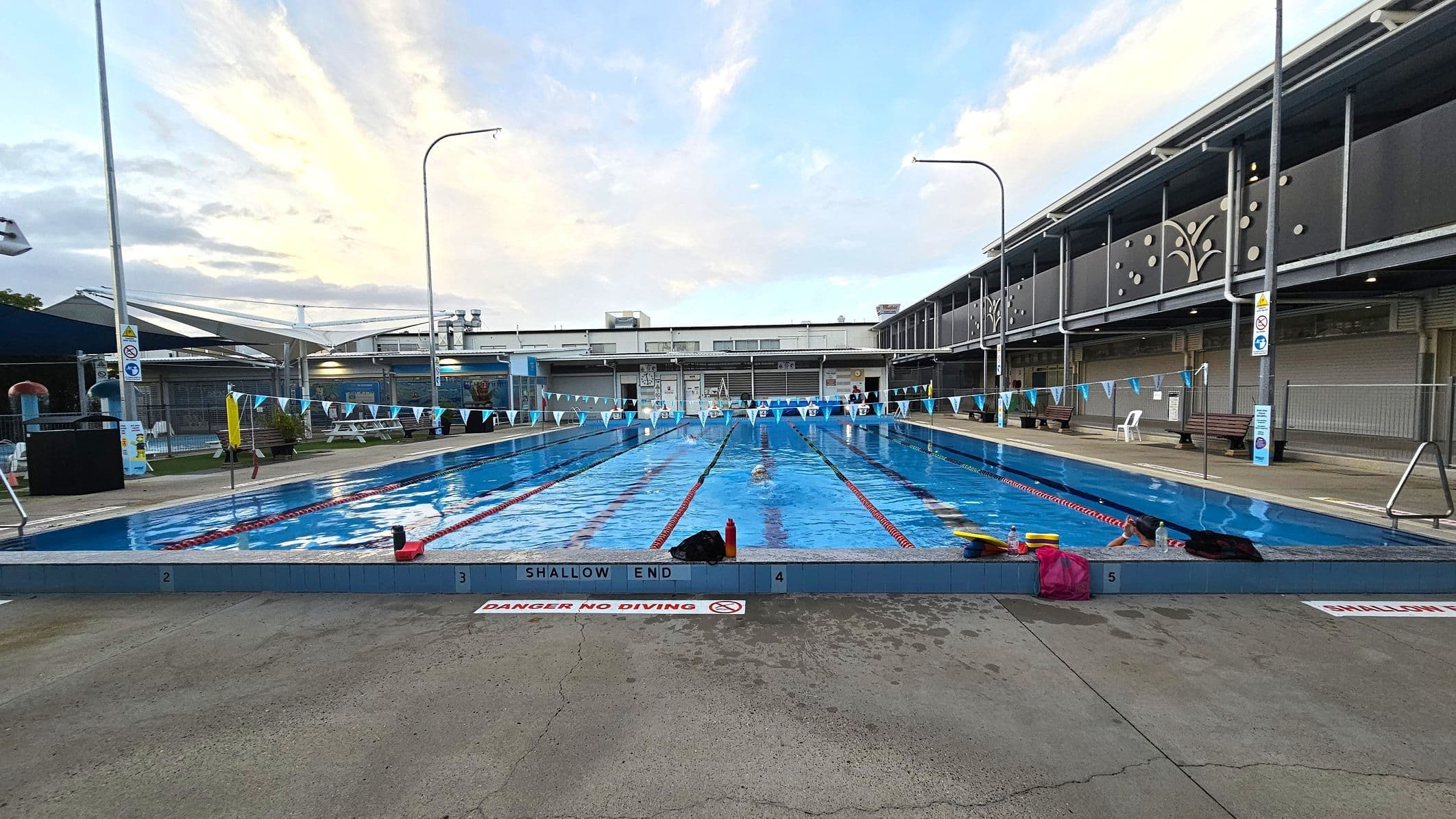 An outdoor swimming pool with multiple lanes, each marked by lane ropes. The pool is surrounded by a concrete deck with a "Caution Deep Water 1.5m" sign. In the background, there is a two-storey building with decorative panels and large windows. The building has a covered walkway on the upper level. Poolside benches and a few safety cones are visible. The area is well-lit with tall lights, and a few people are swimming in the lanes.