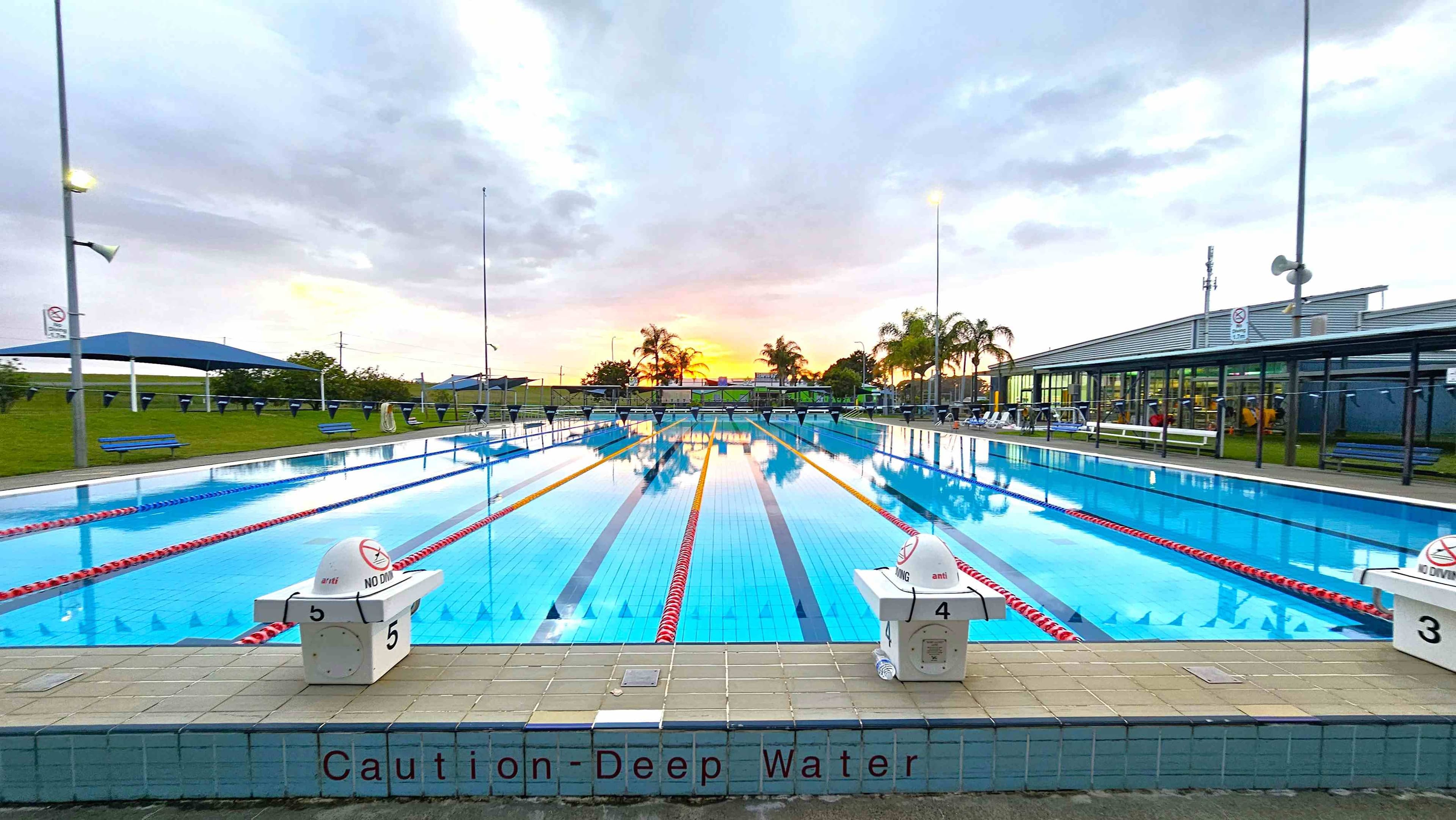 An outdoor swimming pool with marked lanes stretches into the distance. On the left and right, there are diving blocks labelled with numbers and "No Diving" signs. The pool is surrounded by a tiled deck with a "Caution - Deep Water" warning. In the background, palm trees and a building with large windows are visible. The sky is overcast with a hint of sunset colours. Benches and shaded areas are on the left, and a grassy area is visible beyond the pool.