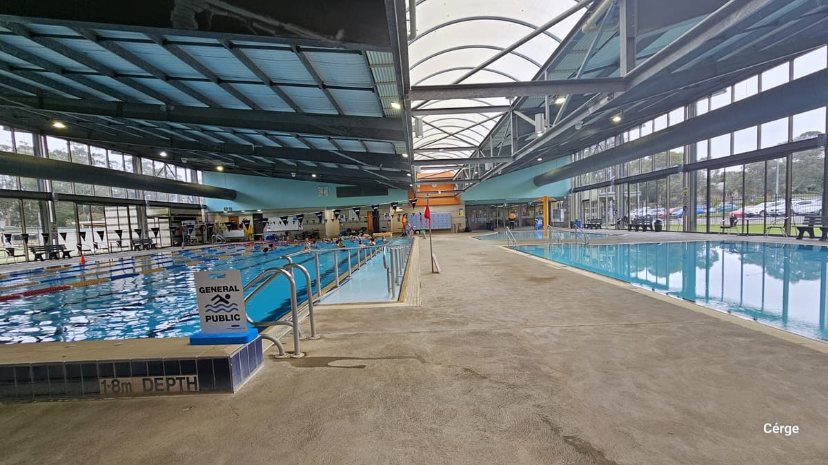 Yarra Aquatic Centre showcases a high-ceilinged pool area with visible pipes and steel beams. A toddler beach pool is positioned in front. In the middle, a yellow steel structure creates a shower or fountain effect. On the left, a larger pool with an entrance ramp and handrails is occupied by swimmers. The locker area is on the right. Bench chairs against the wall at the far end provide seating. The glass window offers a view of the grassy outside.