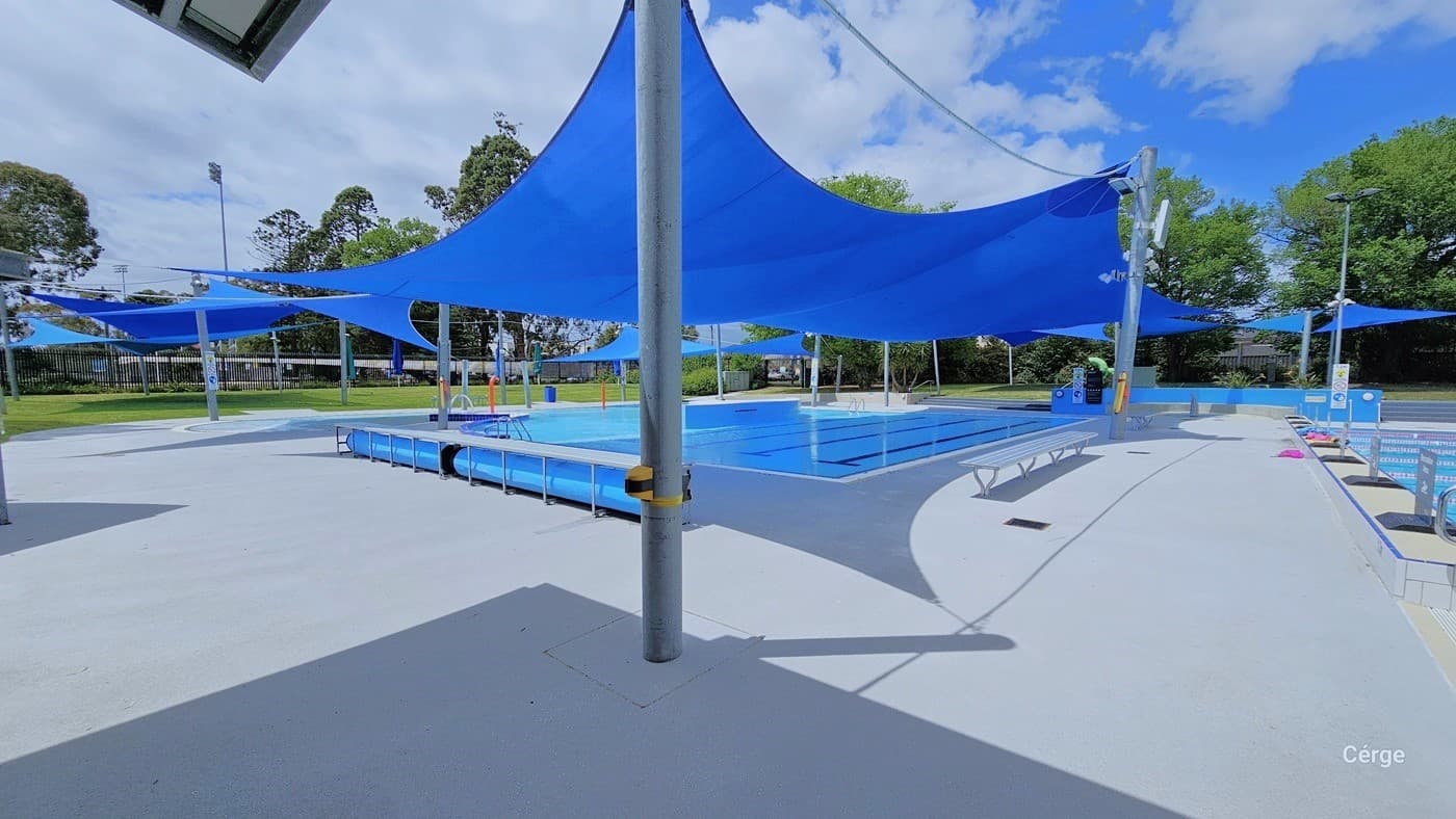 Bird’s eye view of the Werribee Outdoor Pools. Over the heated leisure pool are large sun shade sails wrapped around the grey metal posts. Some of the posts have white signs for the pool rules. Noticeable on the bottom of the outdoor pools is the bold black line that forms a T at each end to alert swimmers that the end of the pool is approaching. White benches are available throughout the area. From this image, the facility has a semi-circular grey corrugated roof.