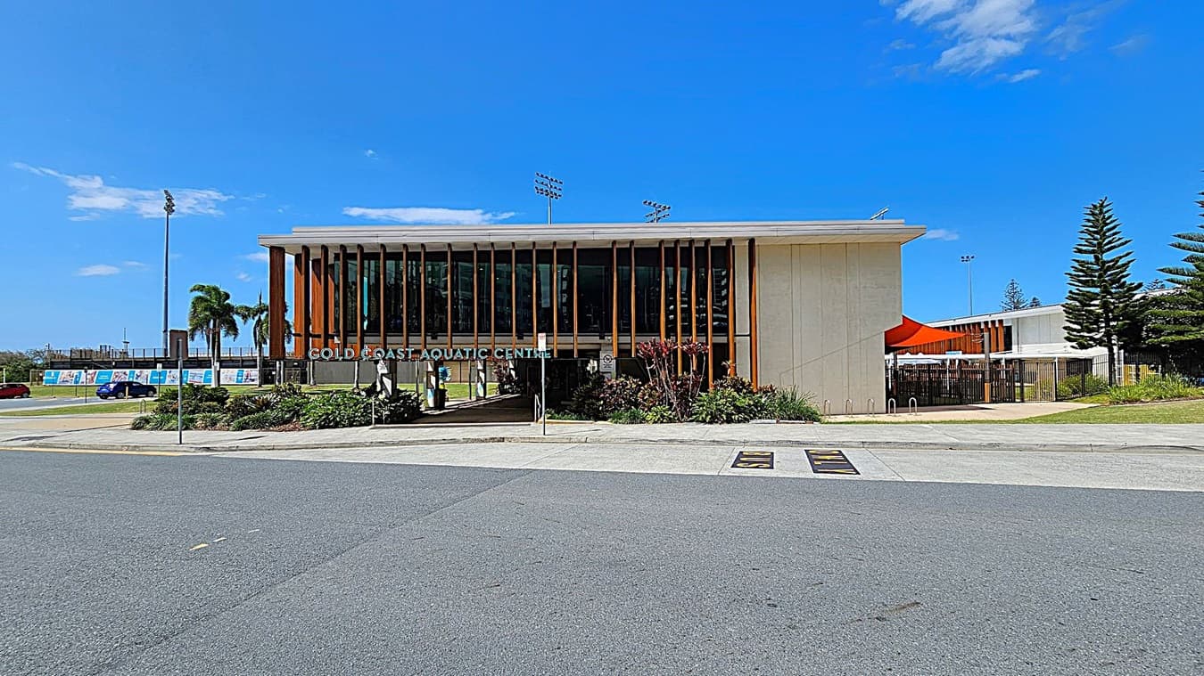 A modern building with a flat roof and large vertical wooden slats on the facade. The sign reads "Gold Coast Aquatic Centre." In front, there are landscaped gardens with various plants. To the right, an orange canopy is visible, and there are tall pine trees. The road in the foreground is wide and grey, with a pedestrian crossing marked. In the background, there are palm trees and a few parked cars.