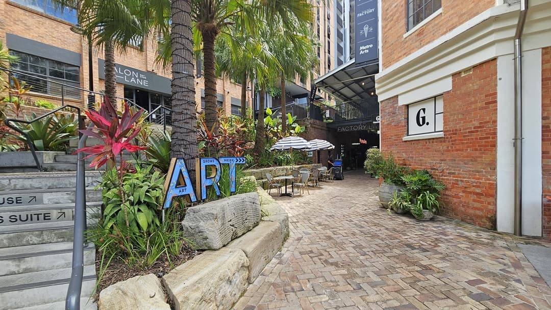 A stairway with handrails leads to a brick-faced building. Plants and palm trees adorn the front, with an "ART" sign. To the right stands another brick building marked "G" with directional signs. On its left, large striped umbrellas shade arranged tables and chairs. Further back, a shaded area with ornamental plants against the wall. The brick floor extends to the rear.