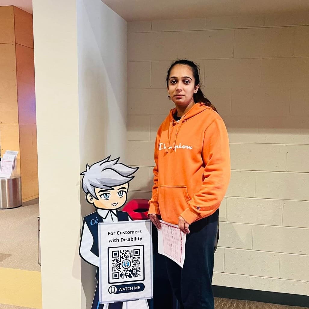 A woman in an orange hoodie stands next to a Cérge cartoon cutout with a QR sign.
