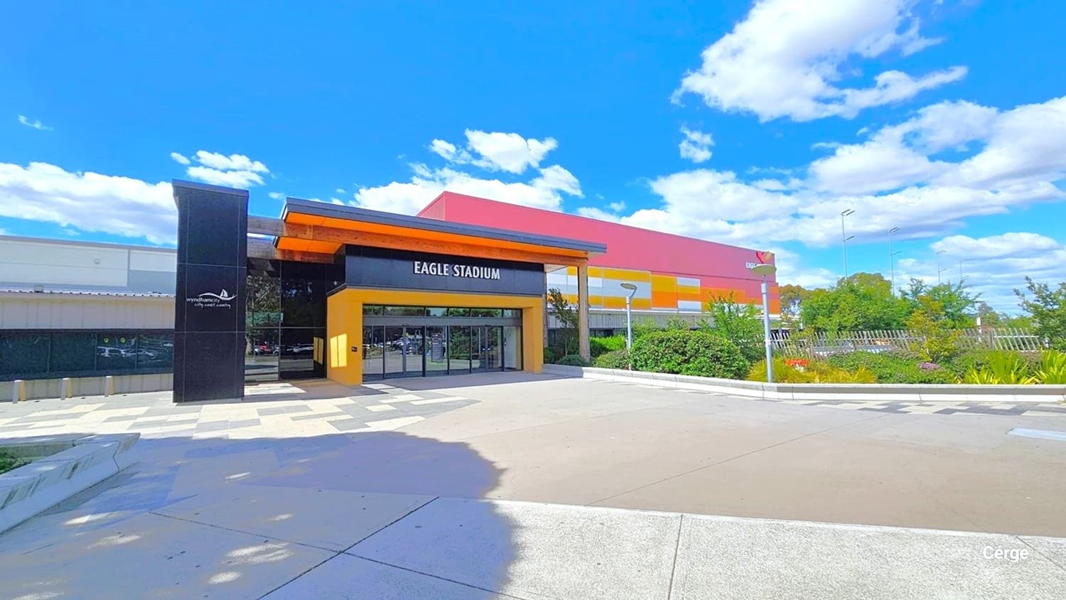 Eagle Stadium concourse with different floor styles. On either side of the concourse are alternating dark and light grey tiles, while in the middle is a smooth cemented floor connected to the automatic, glass sliding doors with black metal frames. On the left side of the floor-to-ceiling glass window is a black tile building column with the Wyndham City logo and slogan: city.coast.country. The name of the facility is detailed on the black wall above the entrance doors.