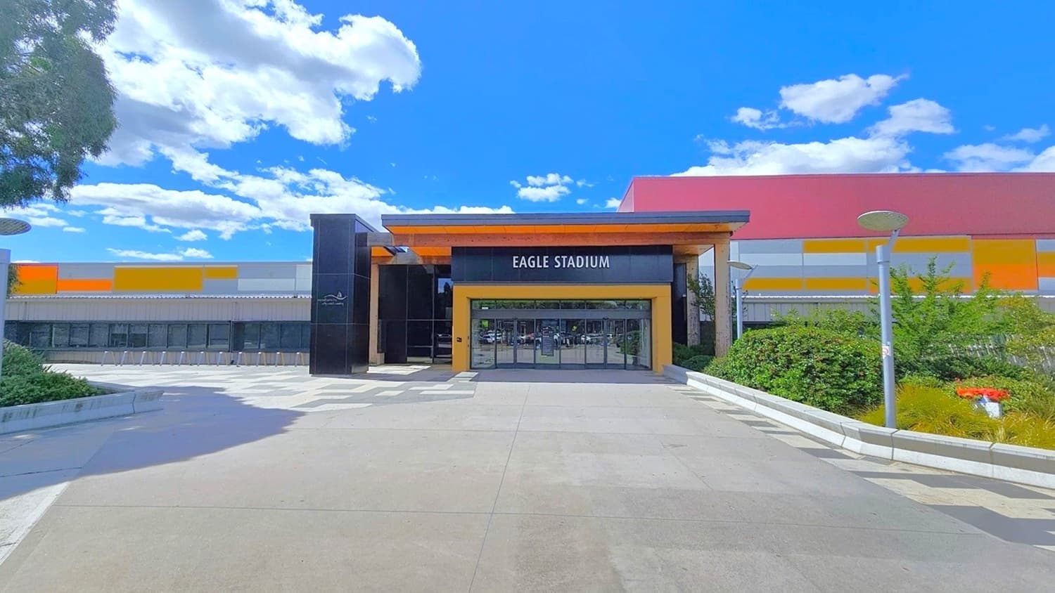 Front view of the automatic glass sliding doors with black metal frames. On the left of the entrance, in front of the grey corrugated wall with glass windows in the middle, are bike racks. The concourse will be well-illuminated by outdoor lamp posts with circular flat heads lined near the border of the garden on the right side.