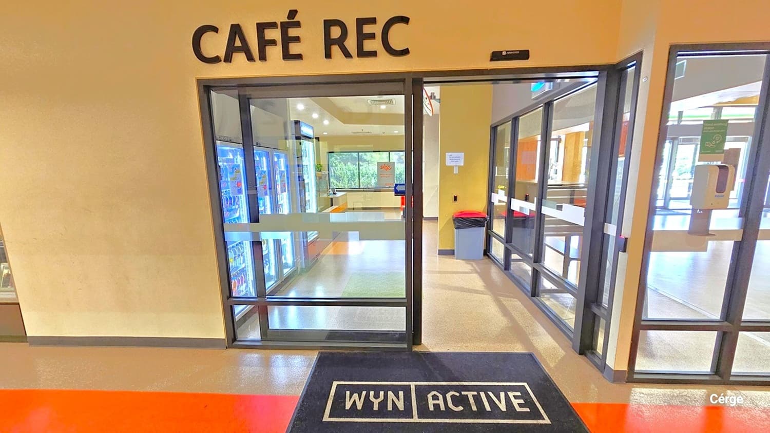 Café REC at Eagle Stadium has automatic, glass sliding doors with black metal frames. The Café also has glass windows, which allow a view of the reception area and entrance. In front of the Café door is a WYN ACTIVE black mat placed on top of the orange-painted floor. On the glass window to the left is a sanitizing dispenser. There is a grey garbage bag with an orange lid in front of the fridge.