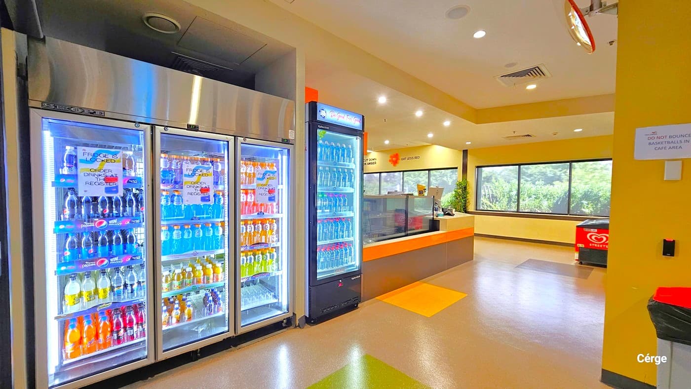 Multi-tiered white fridge with silver stainless steel on top. On the yellow-painted column behind the garbage bin with an orange lid is a piece of paper inhibiting the bouncing of basketballs in the Café area. The brown floor has yellow and green square accent tiles. There is an ice cream fridge with the Selecta logo placed against the glass windows.