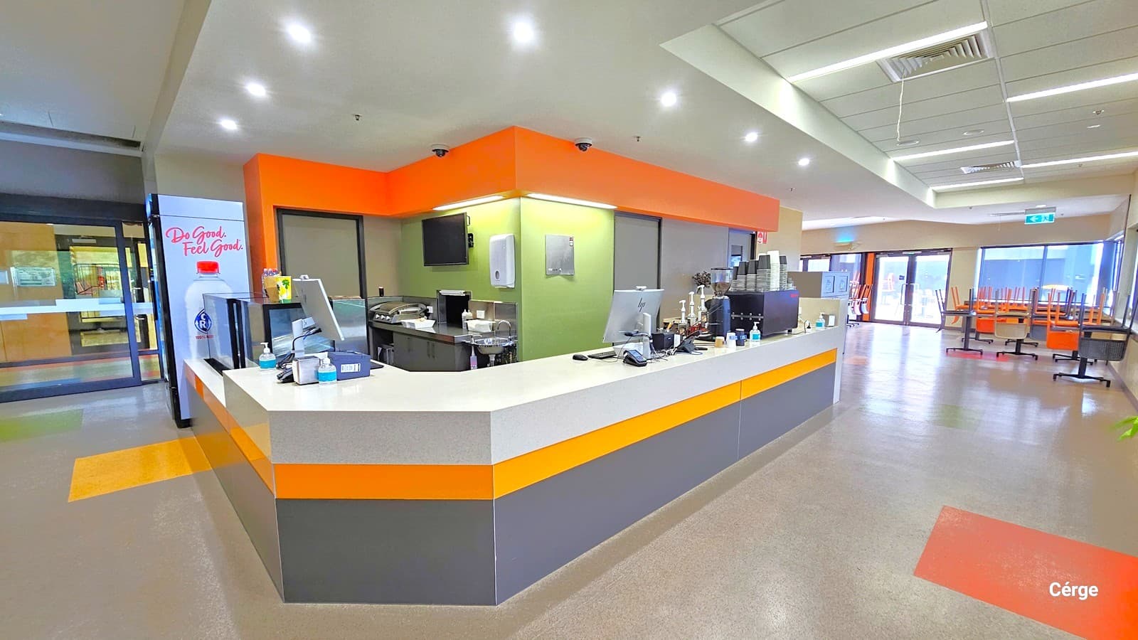 The Café has a light grey countertop with dark grey walls and yellow between them. There are two grey doors with black door outlines behind the countertop. The back wall is lime green and has a screen monitor, a sanitizing dispenser, and two CCTV cameras pointing on each side of the Café. Turning left after the entrance are dark grey tables with orange, black, and light brown plastic chairs.