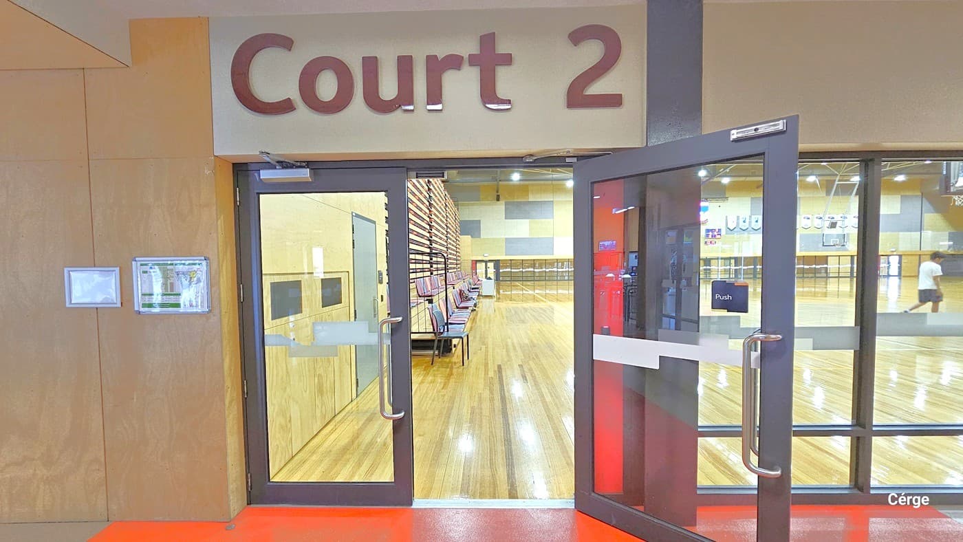 Entrance to a sports hall labelled "Court 2" with glass double doors. The floor is wooden, and the ceiling has visible lighting fixtures. Inside, there are benches along the left wall and a person visible in the distance on the right. The walls are a mix of wood panelling and light colours. The foreground has a red floor leading into the court.