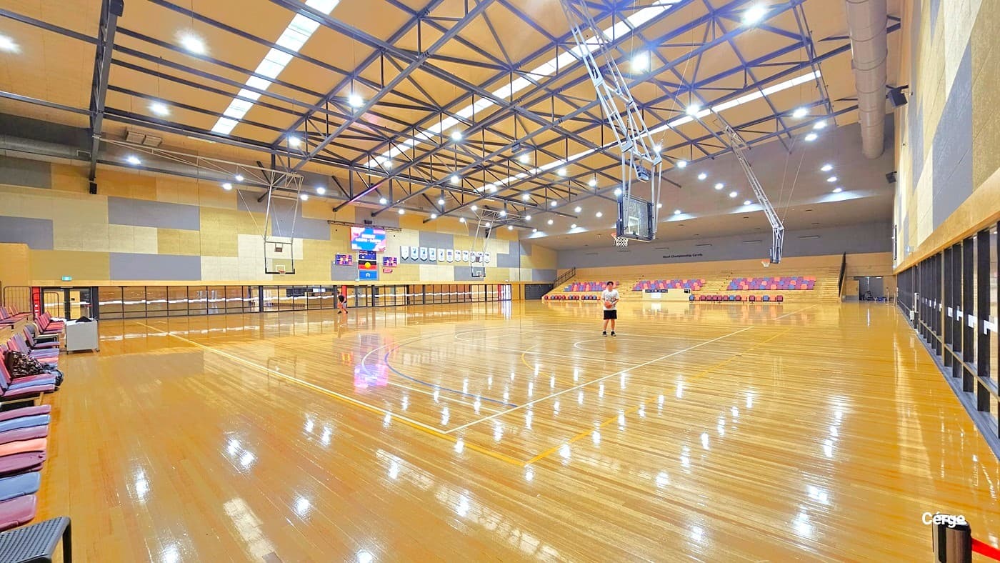 A spacious indoor basketball court with a polished wooden floor. On the left, there are rows of seating with a few people sitting. In the centre, a person stands on the court, and a large scoreboard is visible on the far wall. The right side features more seating and a series of windows. The ceiling is high with exposed beams and bright lighting.