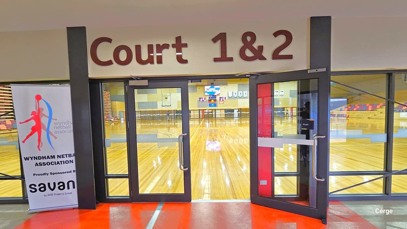 Glass doors leading to a sports hall with a wooden floor, visible through the doors. Above the entrance, a sign reads "Court 1 & 2". To the left, a banner for the Wyndham Netball Association is displayed. The floor in the foreground is red and grey. In the background, sports equipment and seating are visible inside the hall.