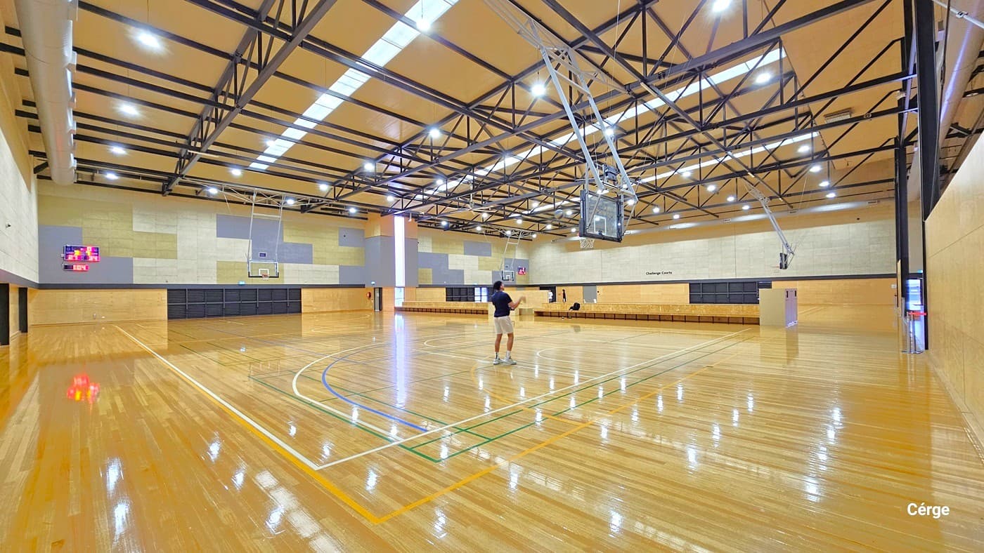 A spacious indoor basketball court with a polished wooden floor. In the centre, a person is standing near the three-point line. The ceiling features exposed metal beams and bright overhead lights. On the left wall, a digital scoreboard is visible. The right side has a series of benches along the wall. Two basketball hoops are suspended from the ceiling, one in the foreground and another in the background. The walls are a mix of light wood and grey panels.