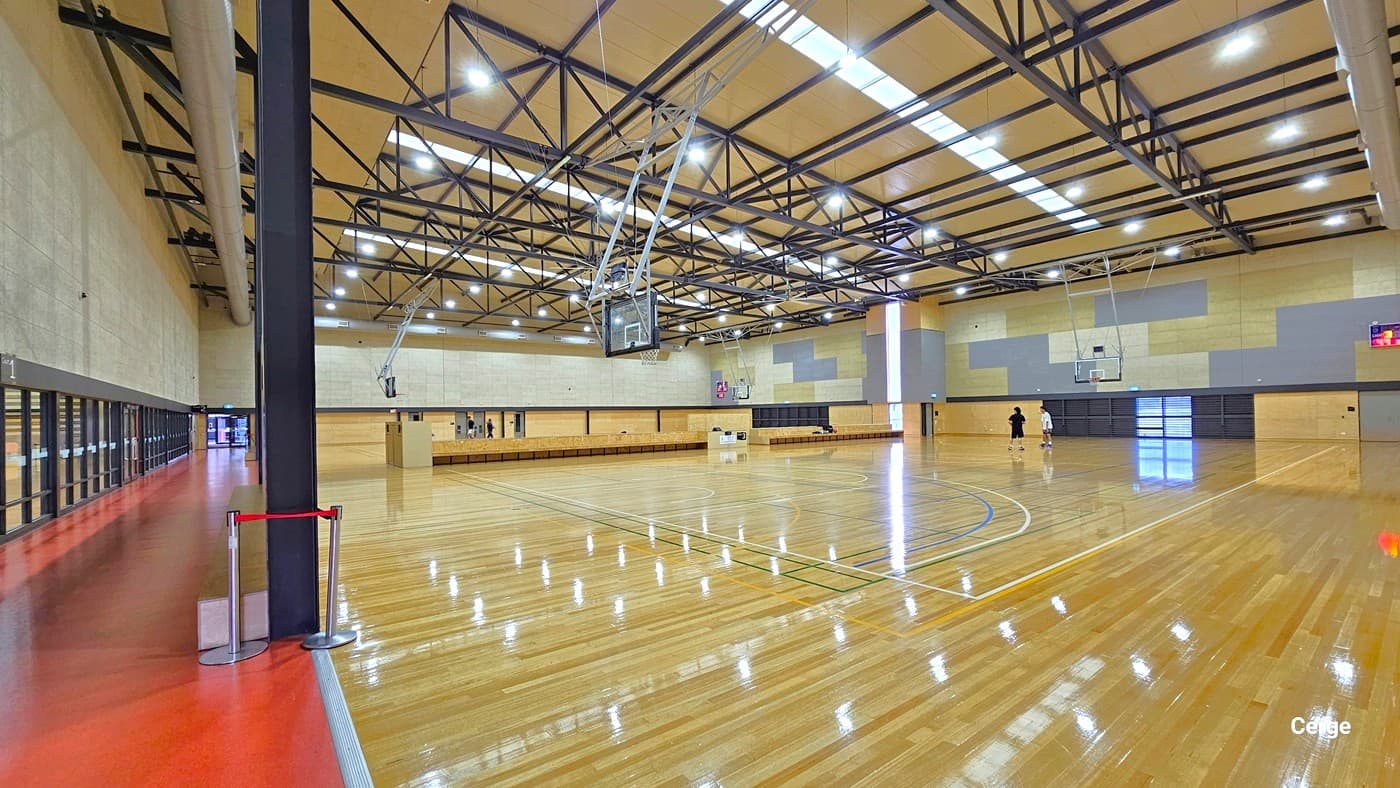 A spacious indoor basketball court with a polished wooden floor. On the left, a red walkway runs alongside a wall with large windows. In the centre, a basketball hoop hangs from the ceiling, which features exposed beams and bright lighting. On the right, two people are on the court near another hoop. The walls are light-coloured with some darker panels. A digital scoreboard is visible on the right wall.