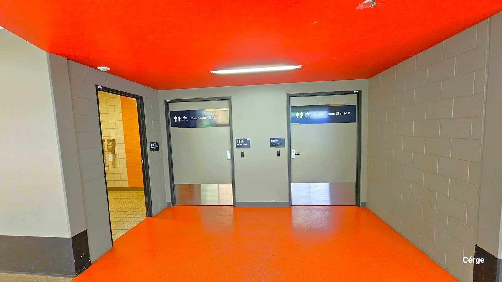 A corridor with three doors. On the left, an open door reveals a tiled room with a water fountain. In the centre and right, two closed doors are labelled "Male Group Change A" and "Male Group Change B" with gender symbols. The floor is bright orange, and the ceiling is red with a fluorescent light fixture. The walls are grey with a block pattern.