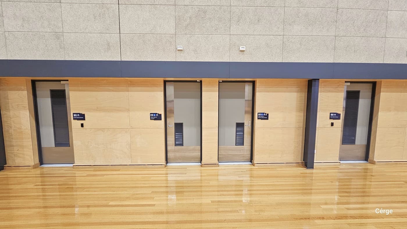 Four doors are aligned side by side on a light-coloured wall. Each door has a small sign with symbols indicating restroom facilities. The floor is polished wood, reflecting light, and the ceiling is not visible. The wall above the doors is a different texture, with a grey strip running horizontally above the doors.