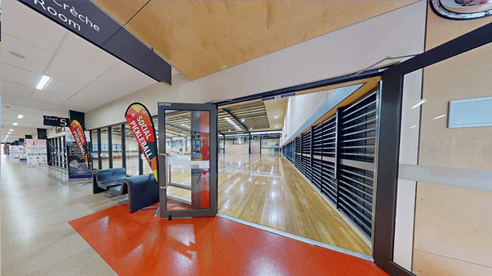 The manually operated glass doors with grey metal door frames are open in Courts 5-8. A rectangular red section is painted on the floor directly in front of the doors. Found within the corridor are large advertisement banners of white, red, and blue. Two unique curvy grey seats are placed left of the door. Beside each seat is a tear-shaped red, orange, yellow, and grey flag that has an X-shaped leg support. There is a large, black metal grating with gaps on the right wall inside the court