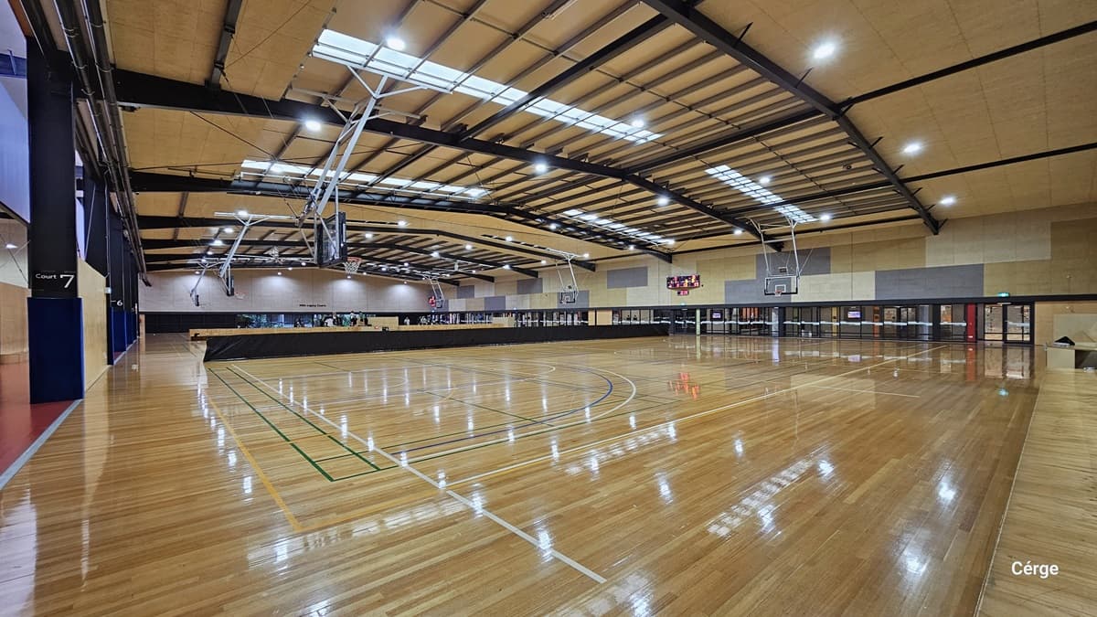 A sign of white Court 7 over a black background is on the left black column near the red-painted floor in Eagle Stadium. The court has polished timber floors with light orange, green, white, and blue lines. The ceiling is brown with a grating-like design of black beams and fluorescent lighting. The upper right walls have an alternating large rectangular tile of dark grey, brown, and light brown. A hip-high, long, black cover separates the courts.
