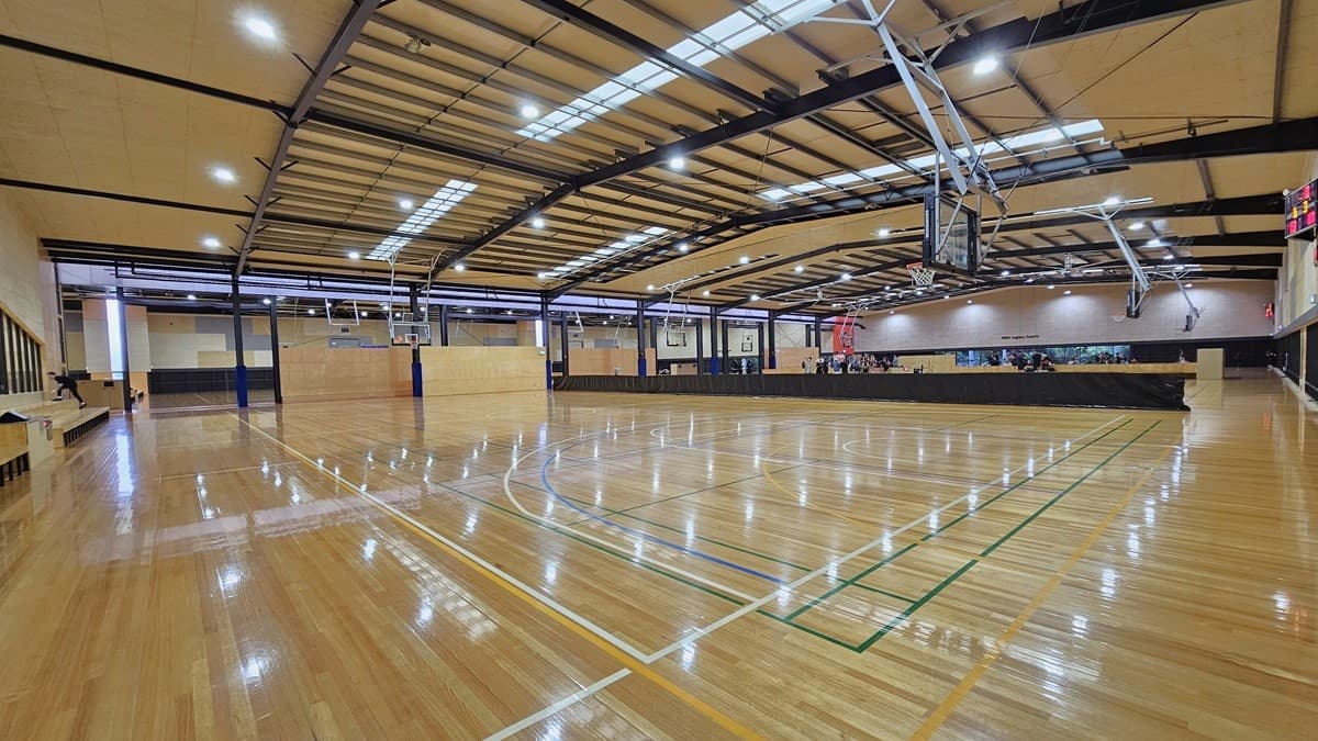 Court inside Eagle Stadium. On the right side is a bench-style seating with long and short black metal beam structures. The court has polished timber floors with light orange, green, white, and blue lines. The ceiling is brown with a grating-like design of black beams and fluorescent lighting. The back walls have alternating large rectangular tiles of dark grey, brown, and light brown. A hip-high, long, black cover separates the courts.