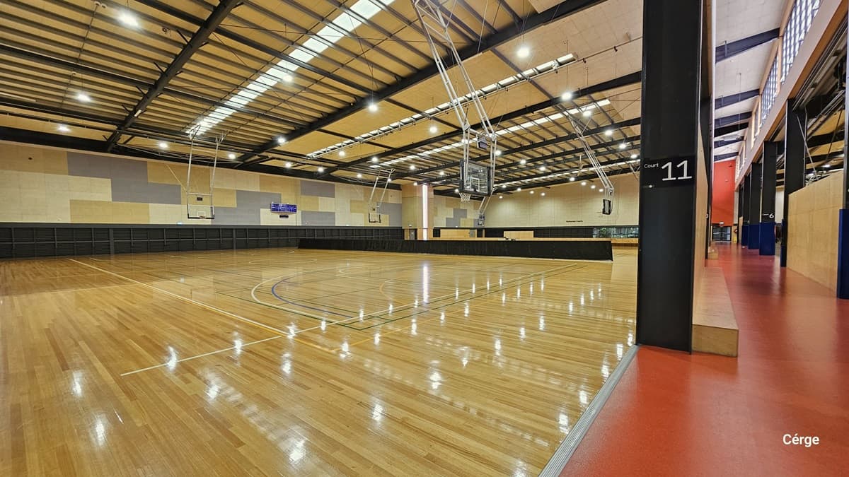 A sign of white Court 11 over a black background is on the right black column with a rectangular brown wood behind near the red-painted floor in Eagle Stadium. The court has polished timber floors with light orange, green, white, and blue lines. The ceiling is brown with a grating-like design of black beams and fluorescent lighting. The back walls have alternating large rectangular tiles of dark grey, brown, and light brown. A hip-high, long, black cover separates the courts.