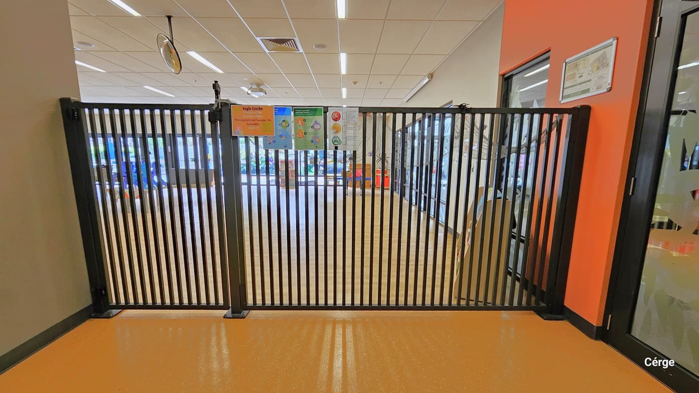 Tall black gates in Crèche on the right side after passing through the entrance glass doors. Small, varying colors of informational signs and notices are placed in the upper part of the gate. A small section of the right wall is orange, while the rest is creme grey. The left wall is also cream grey. There is a small map of the entire facility on the right orange wall.