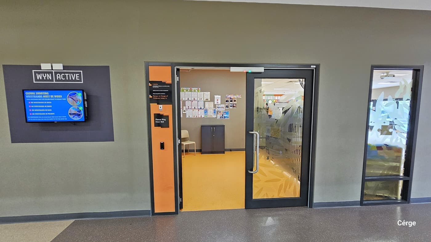 Half-open, manually operated glass doors with black metal frames and curved stainless steel handles of Crèche at Eagle Stadium. The doors have a leaf blur imprint on the glass and are within a wall that has a bottom moss green paint while the upper has white paint. On the right side of the door is a small, narrow orange wall section that has the doorbell. Further to the right is a black square area for WYN ACTIVE and a small monitor screen. The floor is natural grey and light brown stone tile.