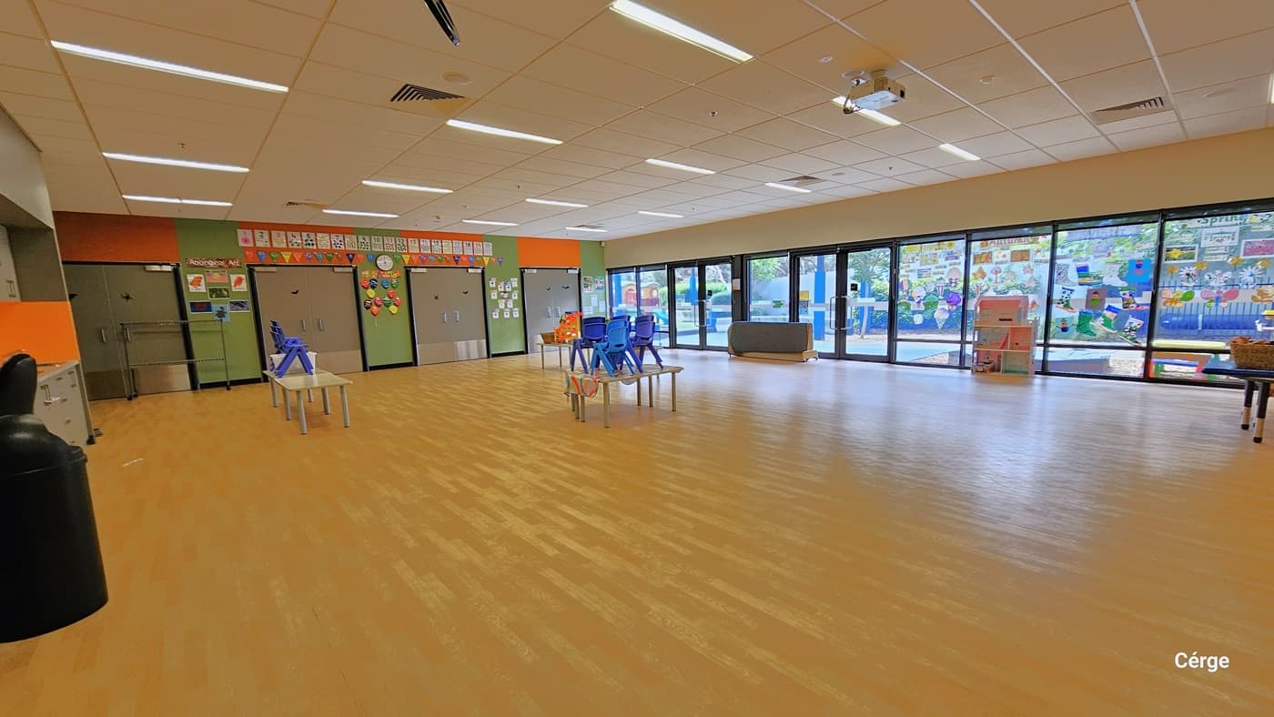 Inside the Crèche at Eagle Stadium. It has light brown timber floors and a white ceiling with fluorescent lights and a white projector mounted in the center. On the left wall is a grey cabinet while on the right wall are glass windows that offer a great view of the enticing outdoor play area for kids. In the middle are small white tables and blue chairs. Colorful interactive designs are placed on the green and orange wall to the back. There are four sets of grey doors to the back.