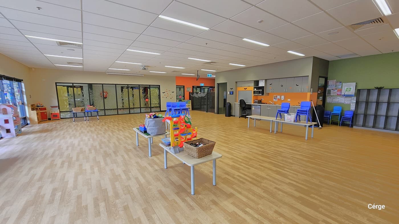 A closer look at the tables and chairs. Above the tables are a grey basket and a netted hyacinth basket filled with varying and colorful toys. Large interactive toys are also available on top of the table. There is a black garbage bin near the entryway and a black office chair on a table near the small kitchen area with modular grey cabinets. A stack of small blue chairs is placed against the right green wall, with several white papers, next to an empty storage rack.