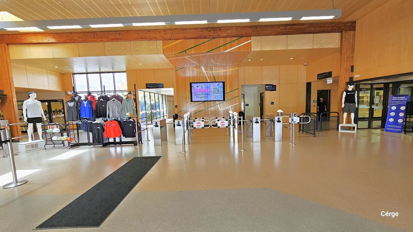 Inside the Eagle Stadium, visitors are greeted by high ceilings that allow for ample natural light. The inside walls are different hues of large wooden brown tiles and a corrugated brown ceiling. The middle wall imitates the court floor with green, white, and yellow lines. To the left is a stanchion post with a black retractable belt that leads to a display of sports apparel, rackets, and balls. Straight ahead are turnstiles that regulate entry, and next to it are black low iron screen gates.