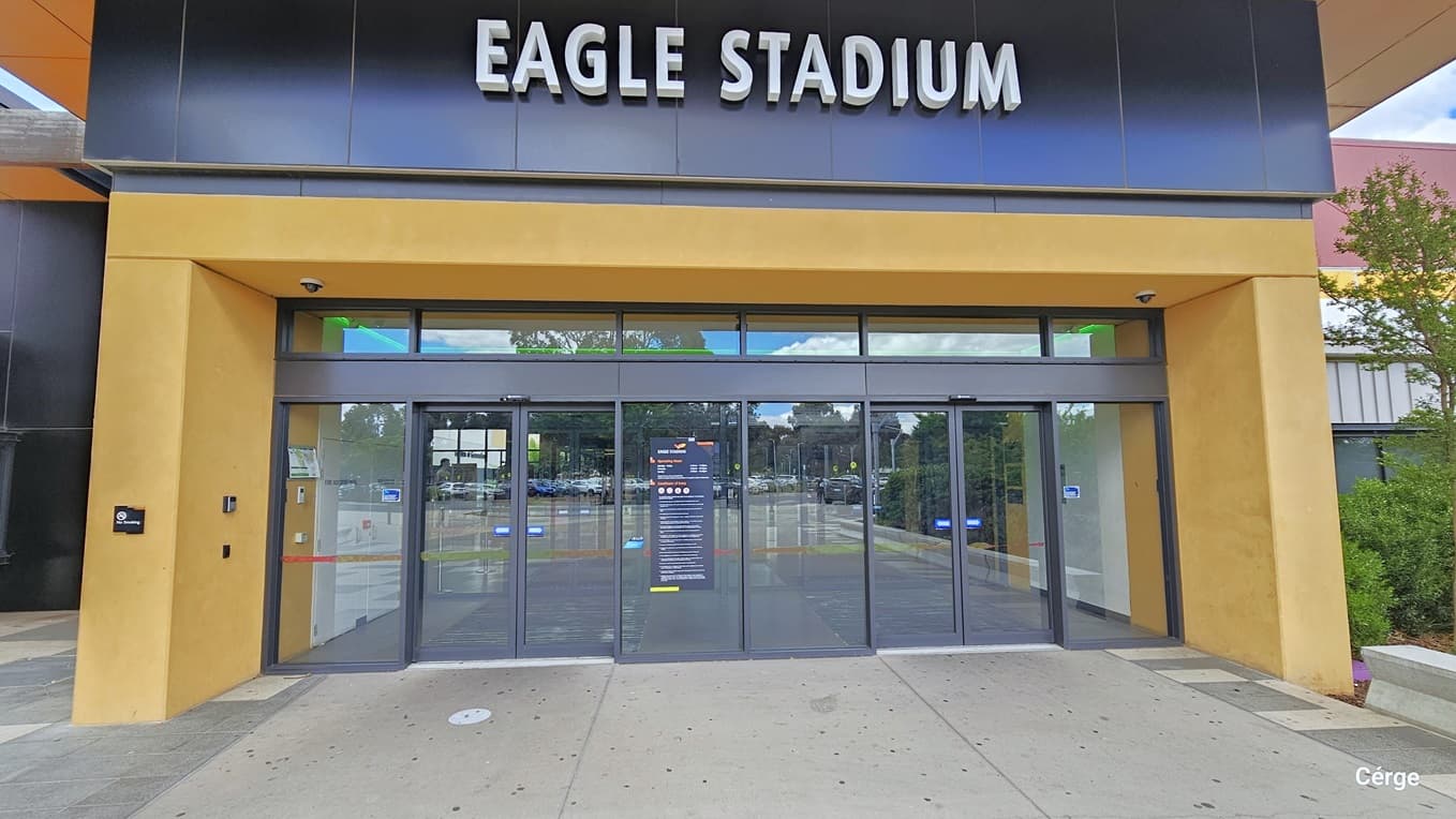 A closer look at the automatic, glass sliding doors with black metal frames of the Eagle Stadium. On the left orange column of the entrance is a black sign for No Smoking. Two CCTV cameras are placed on the left and right side of the ceiling directly on top of the front door. A grey rectangular advertisement signboard of the facility is placed on the middle glass wall. Each sliding door has white arrows with a blue background that are pointing in opposite directions.