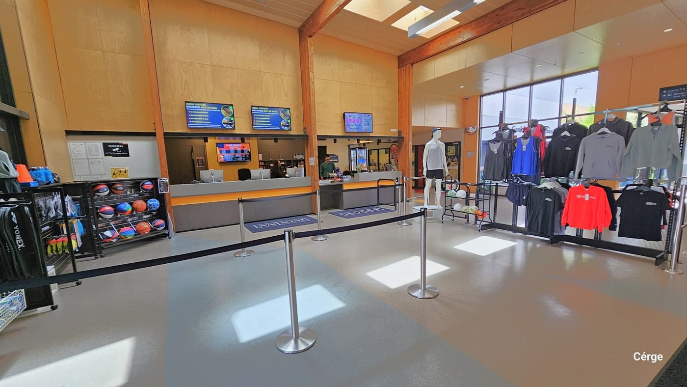 The Eagle Stadium reception area has a grey countertop and wall with an orange accent. On the floor in front of the reception desk are blue mats with WYN and ACTIVE. On the top brown wooden wall of the reception are digital monitor screens. On the left side are black display racks for basketballs, rackets, and tumblers, while on the right is another display for other sports balls and activewear. The guests are regulated by stanchion posts with retractable black belts.