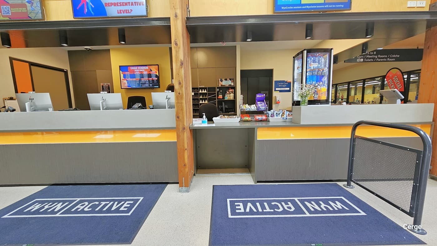 Front of the reception desk are blue mats with WYN and ACTIVE and a grey low iron screen gate. Placed above the grey countertop are snacks and a sanitizing bottle. Further to the right corner, above the counter, is a small fridge with a transparent body for cold drinks. To the left is a glass window. A digital monitor screen is mounted on the yellow wall at the back, between the tall grey cabinets. There is an entry door with a white wall to the right and a blue Center Care Manager sign.