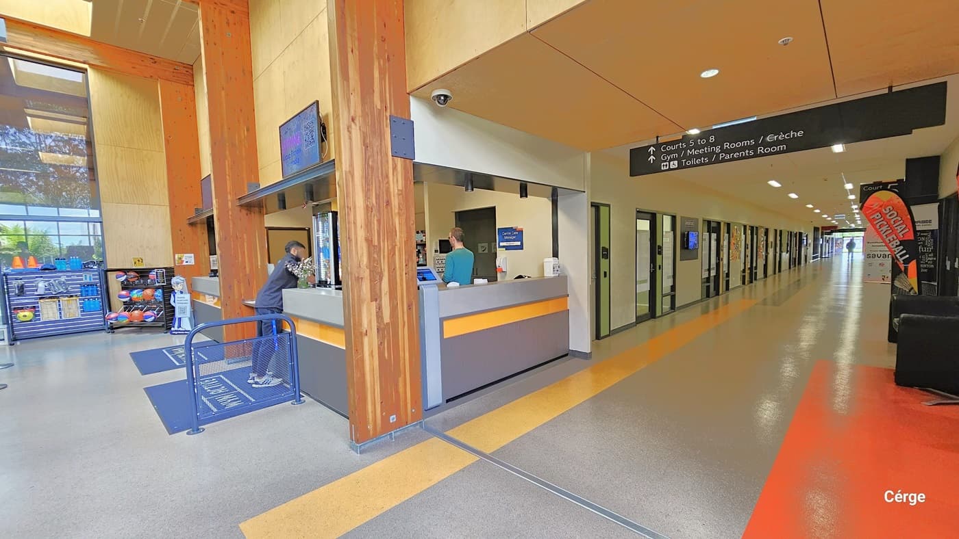 Corridor right next to the reception desk. Image angle shows the Cérge app boy icon holding a QR code beside the black display rack for basketballs in front of the desk. There is a CCTV camera on the light brown ceiling, while in the middle is a black sign for directions to Courts 5 to 8, Gym, Meeting Rooms, and the Toilet and Parents Room. The right wall is white with multiple doors. The floor is grey with a wide yellow line and a large orange floor section on the left.